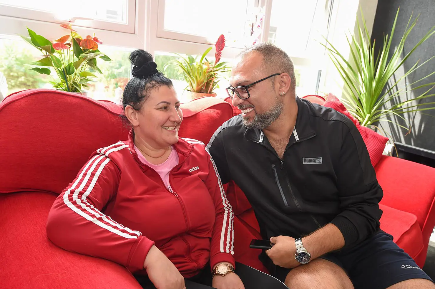A man and a woman sit on a red sofa and smile at each other