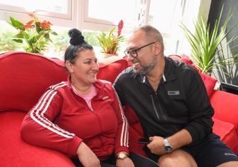 A man and a woman sit on a red sofa and smile at each other