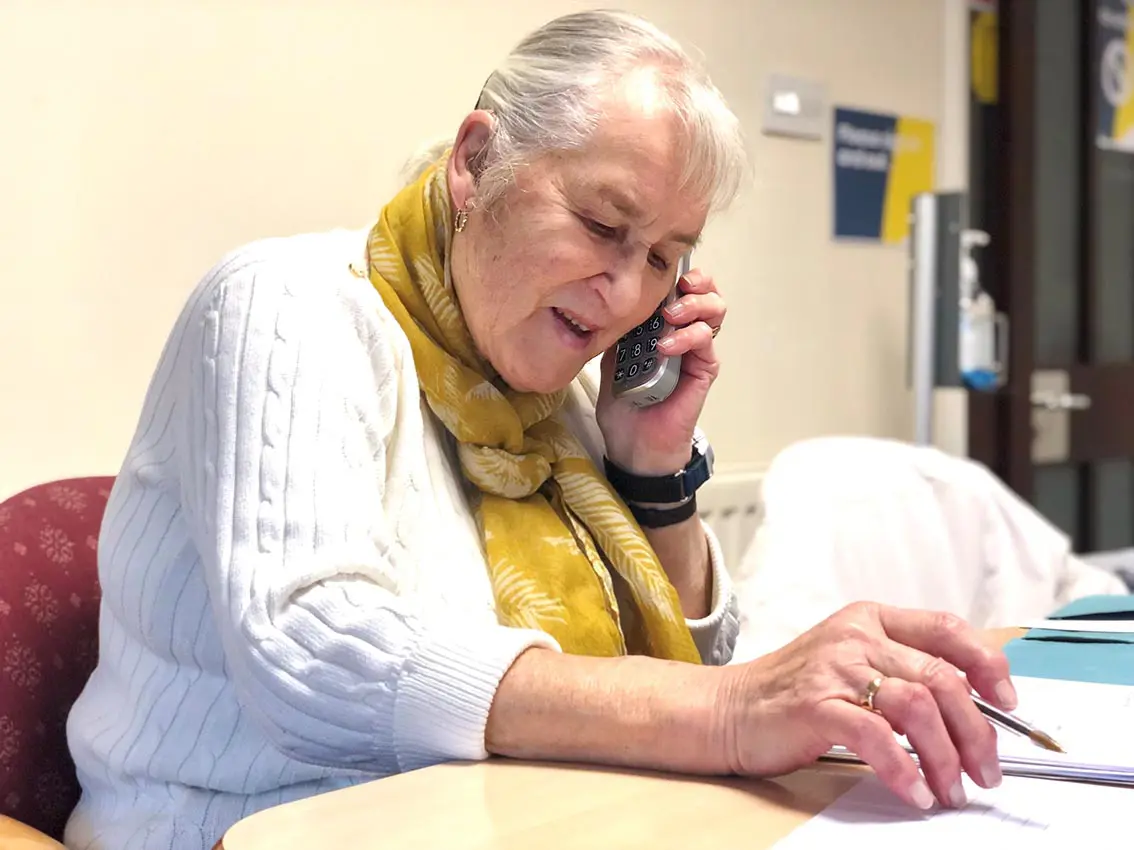 A woman with white hair, wearing a cream cardigan and yellow scarf, talks on a telephone