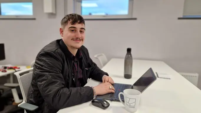 A Wolverhampton Homes apprentice sits at a desk with a mug and laptop
