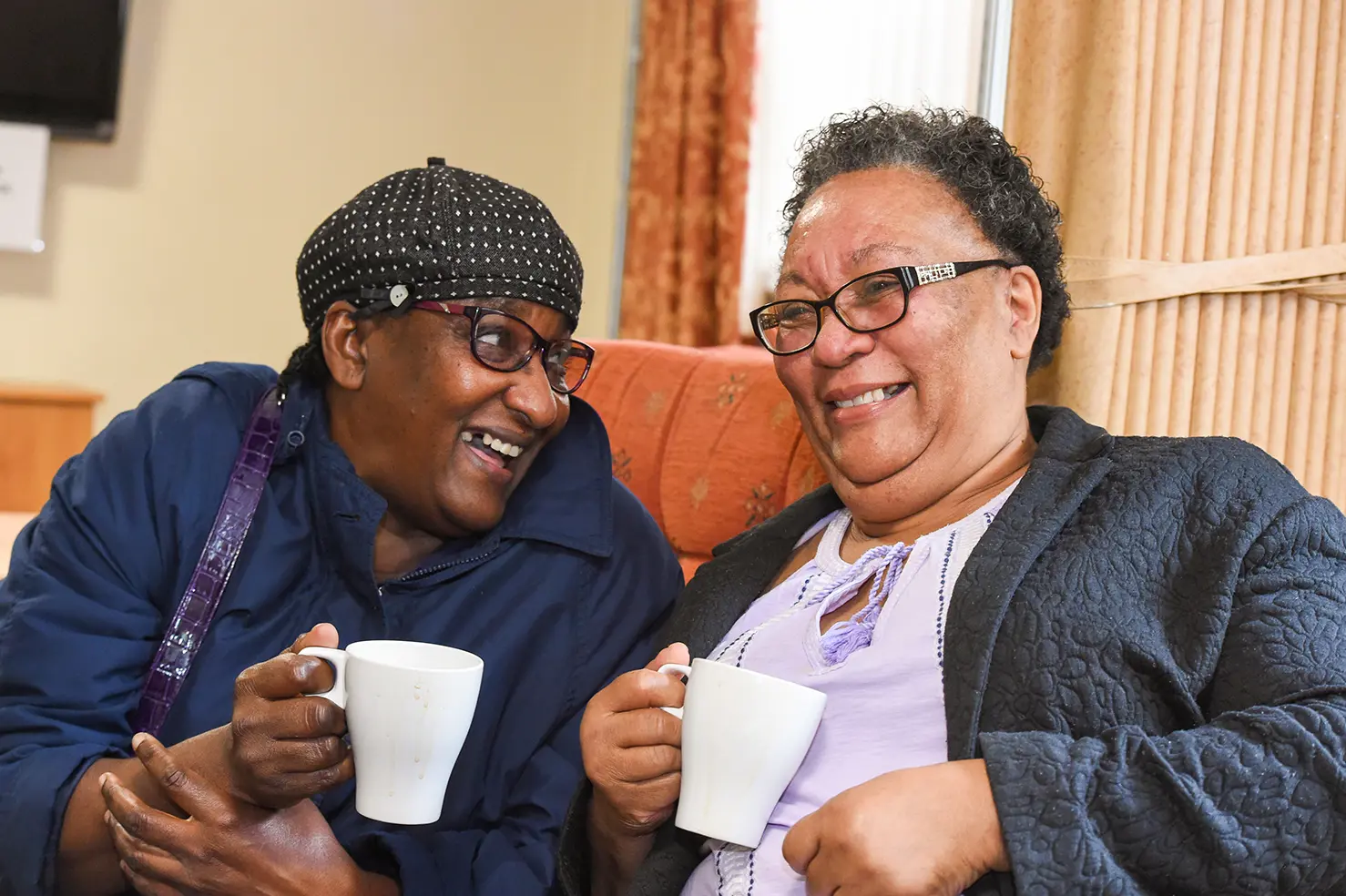 Two women sitting down, both holding mugs and laughing