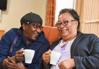 Two women sitting down, both holding mugs and laughing