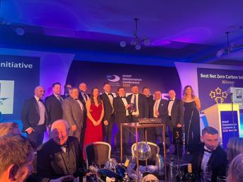 A group of 13 people in suits and evening gowns on stage to accept their award for Best Net Zero Carbon Initiative